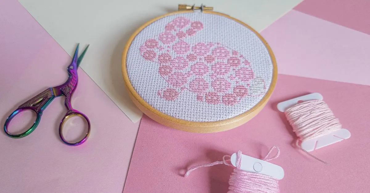 image shows an embroidered easter bunny cross stitch on white fabric framed in a wooden hoop against a pink background with scissors and thread next to it