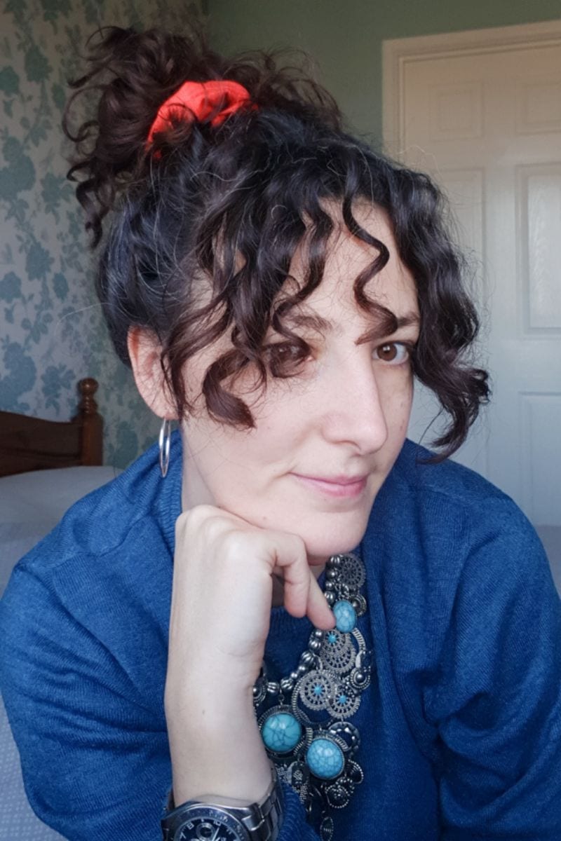 Woman with dark brown curly hair in a pony tail. Resting her chin on her hand looking at the camera