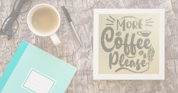 embroidered and framed more coffee please cross stitch next to a coffee cup and notebook
