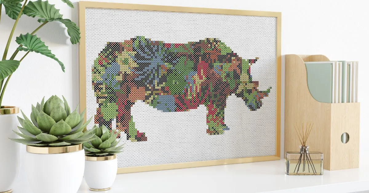 jungle rhino cross stitch design stitched on white fabric and framed in a wooden frame. Positioned on a shelf with plants and ornaments beside it