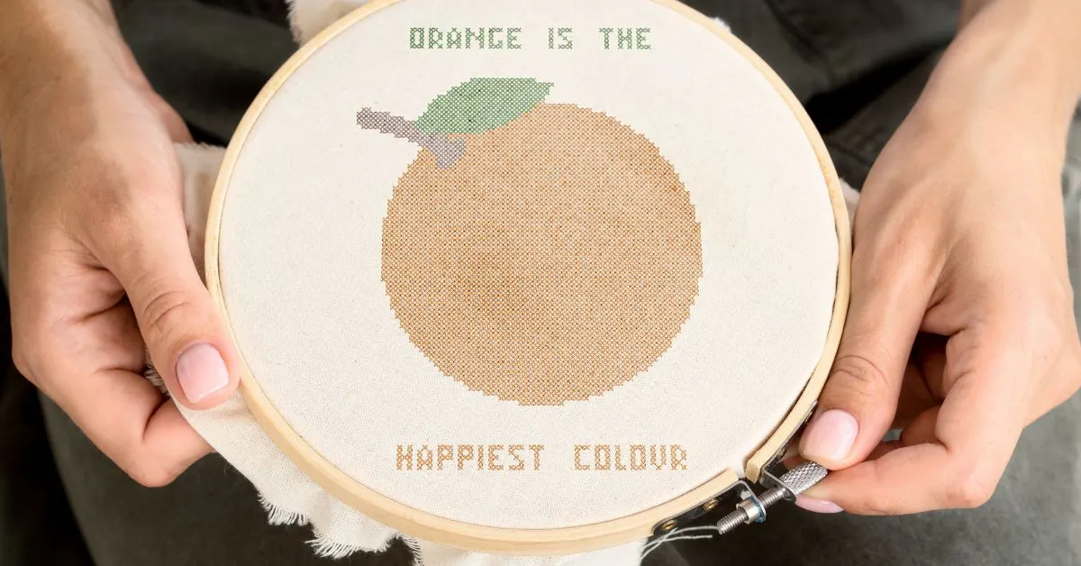 image shows an embroidered orange is the happiest colour cross stitch held in a wooden hoop