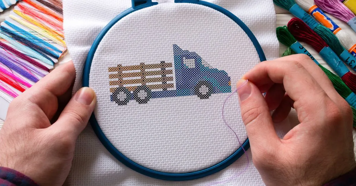 image shows a retro truck being cross stitched onto white fabric in a blue hoop with threads surrounding