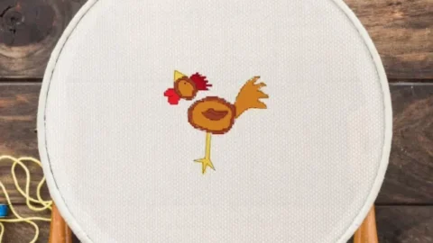 Image shows an embroidered chicken cross stitch framed in a wooden hoop surrounded by sewing tools