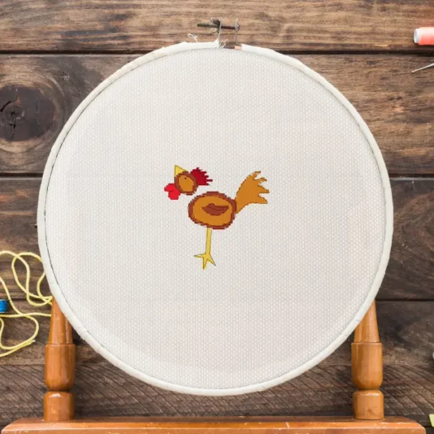 Image shows an embroidered chicken cross stitch framed in a wooden hoop surrounded by sewing tools