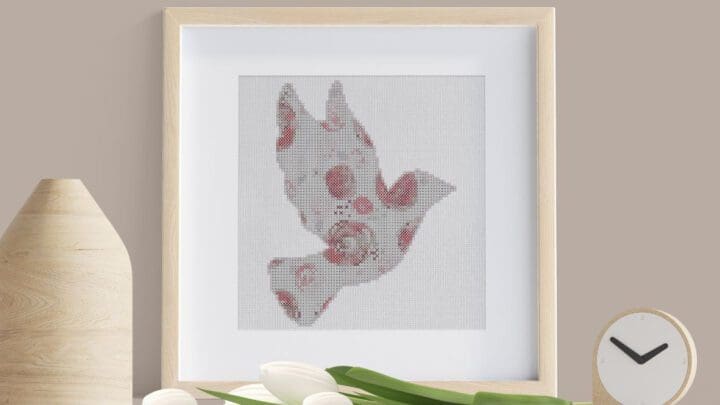 Framed roses dove cross stitch stood on a table with white flowers and a vase.