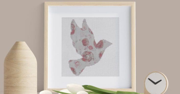 Framed roses dove cross stitch stood on a table with white flowers and a vase.