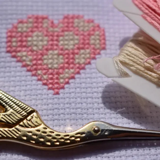 A close-up of a pink and cream cross-stitched heart on white Aida fabric, with embroidery tools nearby. A gold stork embroidery scissors lies in the foreground, while two plastic floss bobbins holding pink and cream embroidery thread are placed beside the stitched design. The image captures the fine details of the stitching and crafting materials.