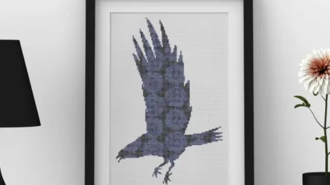 Image shows and embroidered roses raven cross stitch framed in a black frame against a white wall with a black lamp and vase either side of it