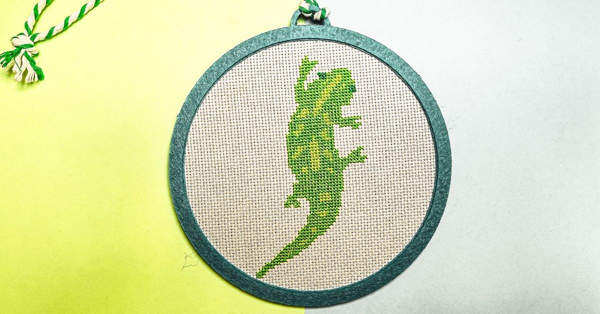framed in a felt hoop stitched gecko cross stitch pattern on a blue and green background