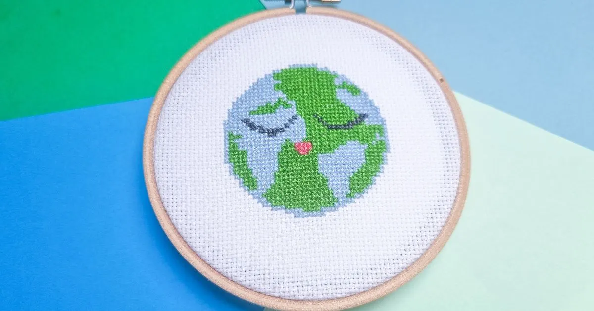 image shows embroidered planet earth cross stitch framed in a wooden hoop against blue and green paper background