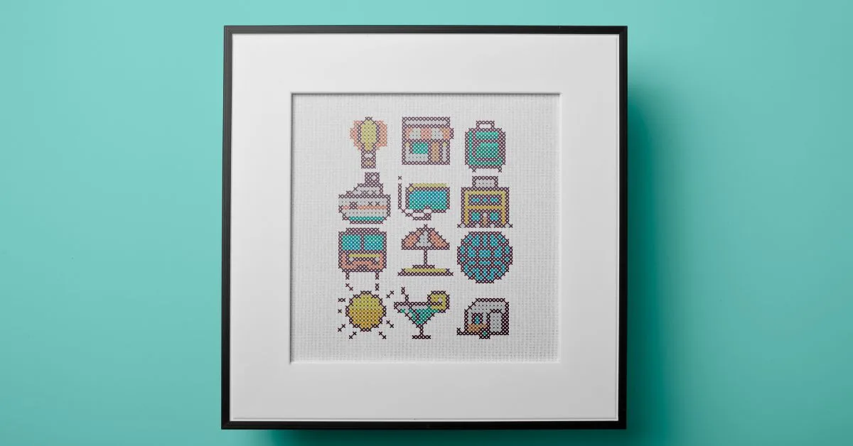 Image shows embroidered 12 tiny cross stitch travel motifs on white fabric framed in a black square frame against a blue wall