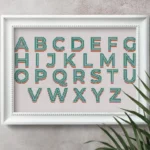 images shows a retro free cross stitch alphabet embroidered and framed in a white frame with a plant next to it