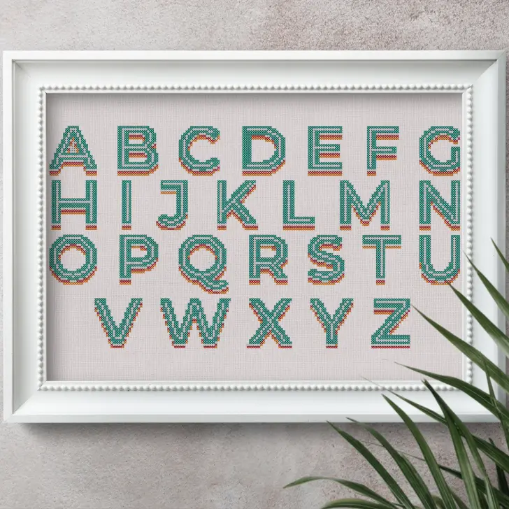 images shows a retro free cross stitch alphabet embroidered and framed in a white frame with a plant next to it