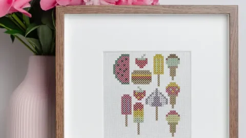 Image shows an embroidered summer treats cross stitch framed in a small wooden frame on a white table with a vase of pink flowers next to it.