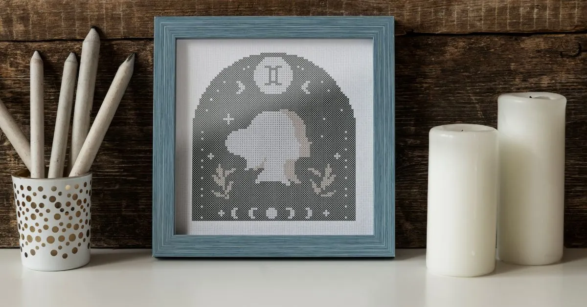 Image shows and embroidered Gemini cross stitch framed in a square blue frame on a white desk with candles and a pot of pencils to the side