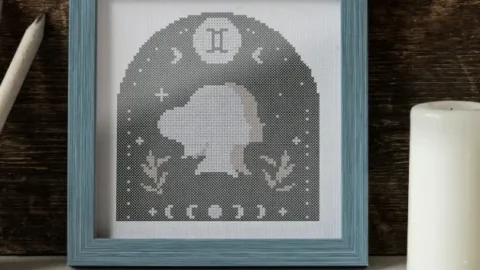 Image shows and embroidered Gemini cross stitch framed in a square blue frame on a white desk with candles and a pot of pencils to the side