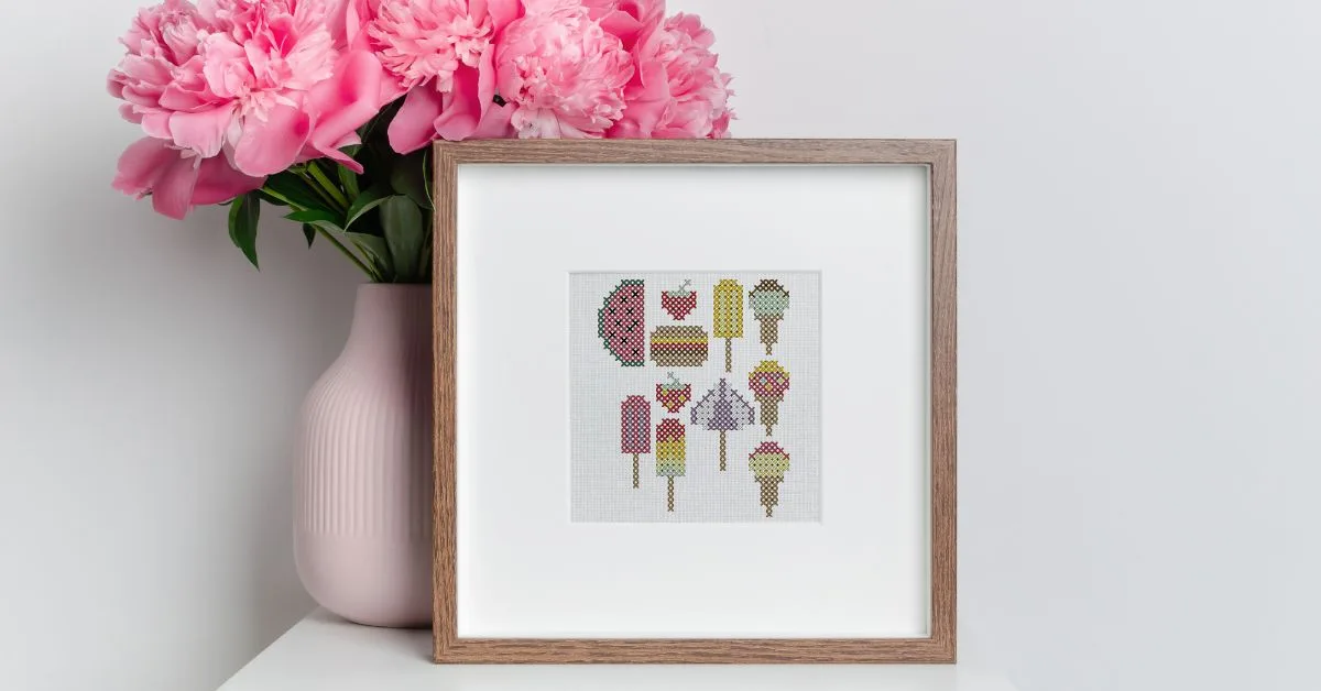 Image shows an embroidered summer treats cross stitch framed in a small wooden frame on a white table with a vase of pink flowers next to it.