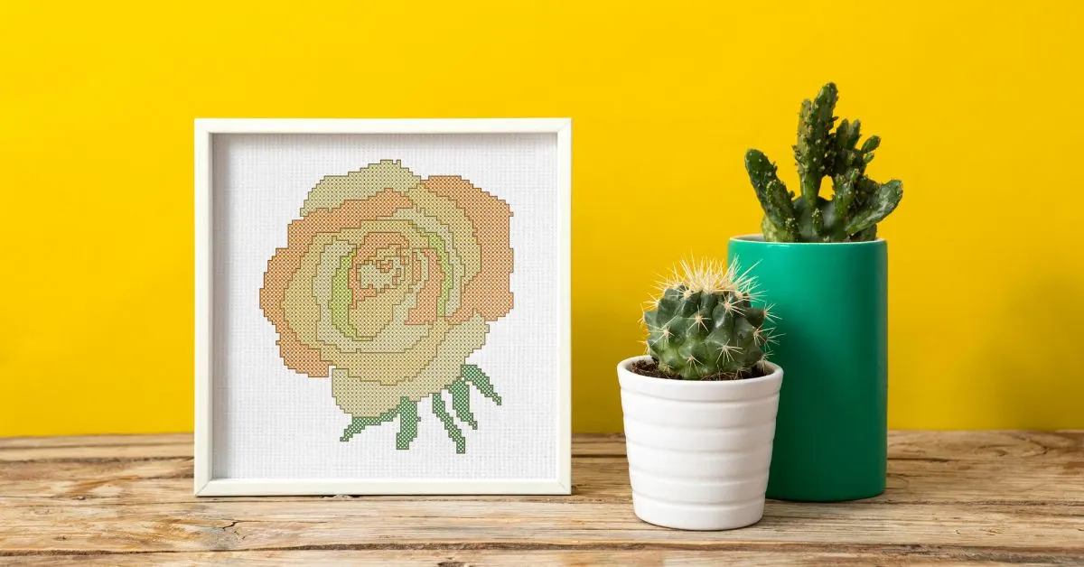 Images shows an embroidered yellow cross stitch rose, framed in a white square frame on a wooden table with two cacti in post next to it