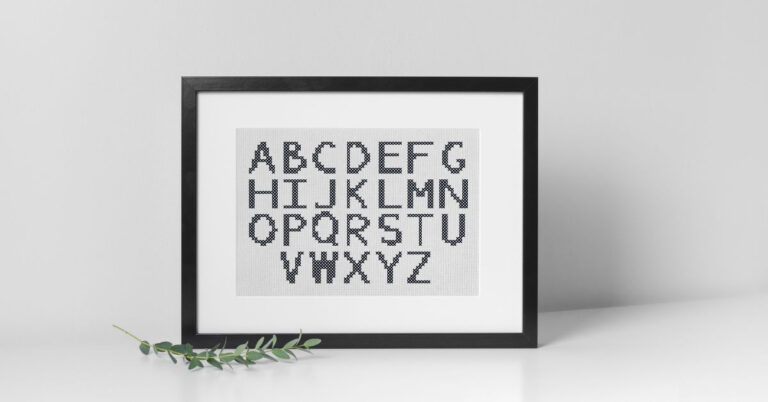 stitched sampler of cros stitch lettering framed in a black landscape frame on a white table