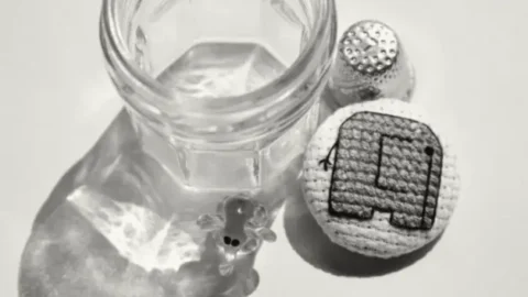 A black and white photograph features a small, empty glass jar with light reflections on its surface, a cross-stitched fabric-covered button displaying a simple mini elephant, a metal thimble, and a tiny glass figurine with black dot eyes. The objects are arranged on a flat surface, casting soft shadows and reflections.