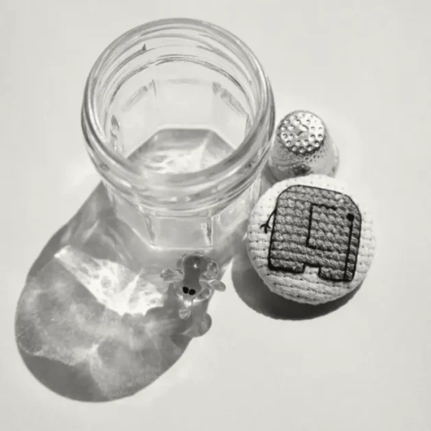 A black and white photograph features a small, empty glass jar with light reflections on its surface, a cross-stitched fabric-covered button displaying a simple mini elephant, a metal thimble, and a tiny glass figurine with black dot eyes. The objects are arranged on a flat surface, casting soft shadows and reflections.
