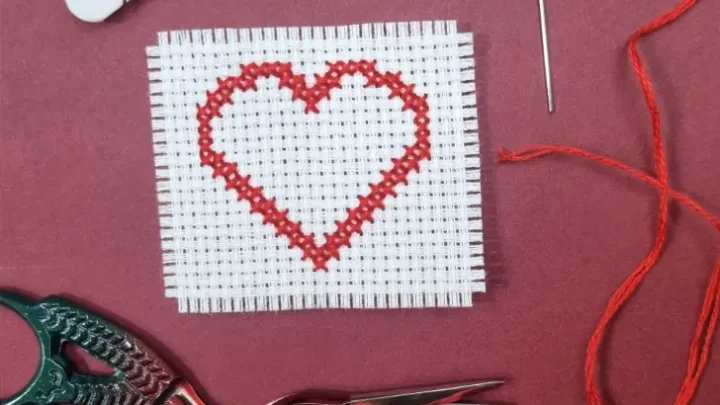 A small cross stitch project featuring a red heart outline stitched on white Aida fabric. Surrounding the fabric are embroidery tools, including a skein of red embroidery floss wound on a plastic bobbin, a needle threaded with red floss, and a pair of ornate stork embroidery scissors. The items are arranged on a deep red background.