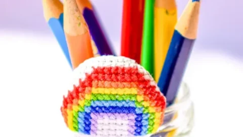 embroidered rainbow cross stitch made into a covered button, decorating a jar of coloured pencils.