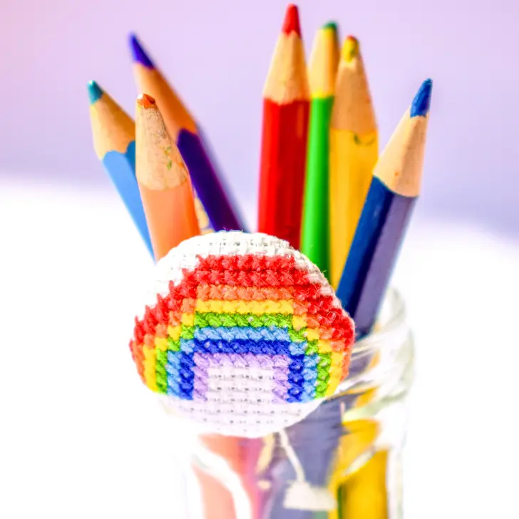 embroidered rainbow cross stitch made into a covered button, decorating a jar of coloured pencils.