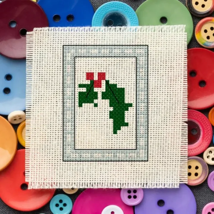 embroidered Free Holly cross stitch pattern on swatch of fabric over a variety of coloured buttons