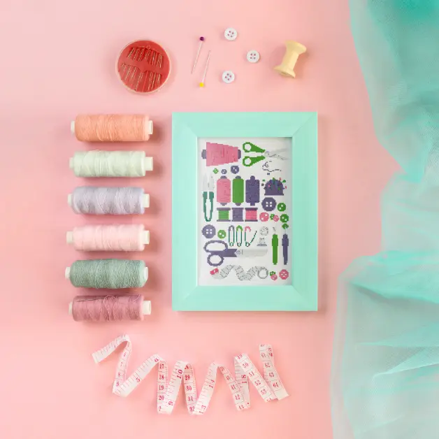framed sewing themed cross stitch sampler surrounded by fabric, thread reels on a pink background