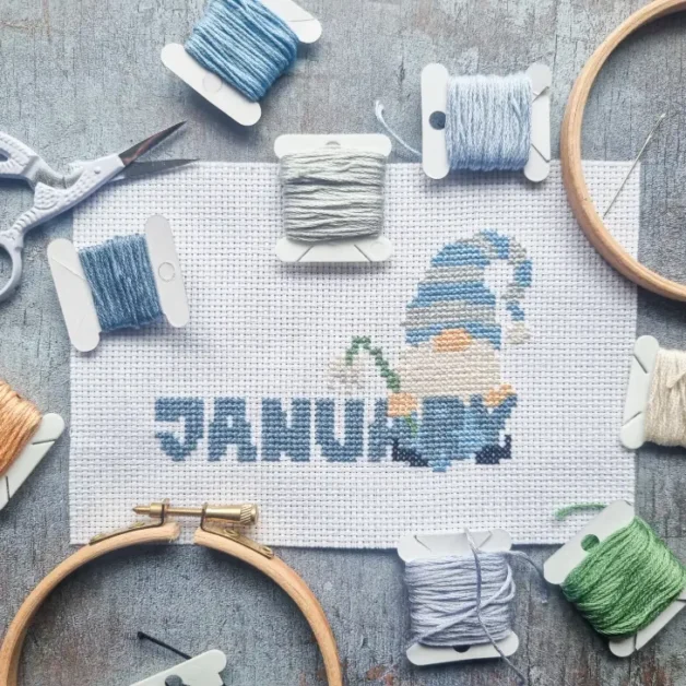 embroidered January gnome cross stitch surrounded by floss bobbins and tools on a blue background