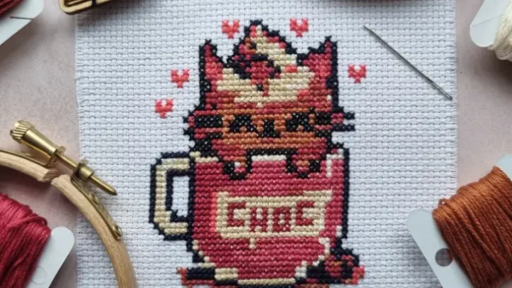 embroidered kitty chocolate cross stitch pattern on a pink background with floss bobbins and scissors surrounding