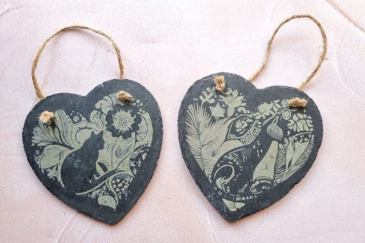 two laser engraved slate hearts with cat designs on them. Left heart features a cat in the garden, right heart features a cat in the wild.