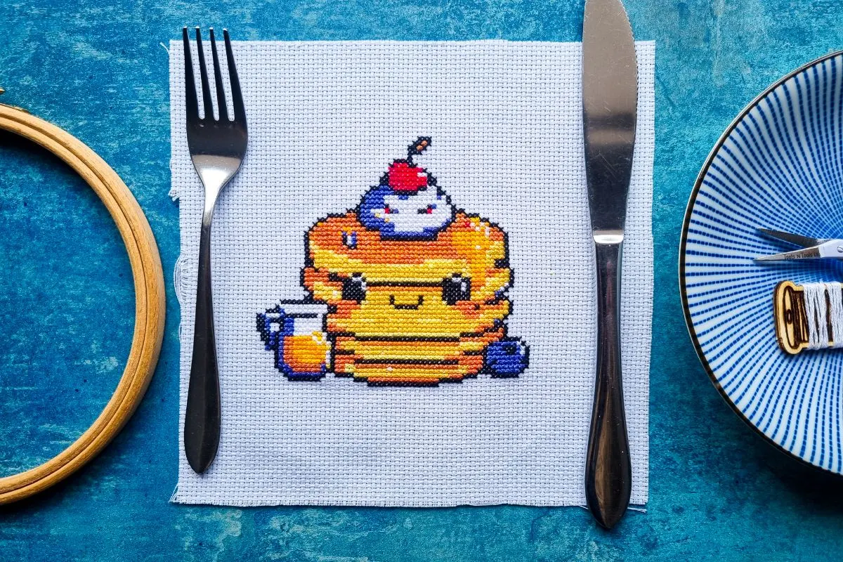 embroidered cute pancake stack cross stitch pattern, with knife and fork wither side on a blue table top