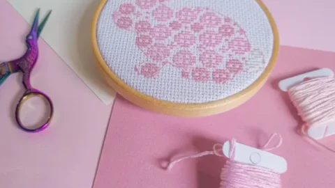 image shows an embroidered easter bunny cross stitch on white fabric framed in a wooden hoop against a pink background with scissors and thread next to it