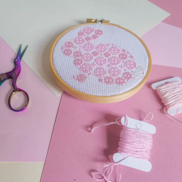image shows an embroidered easter bunny cross stitch on white fabric framed in a wooden hoop against a pink background with scissors and thread next to it