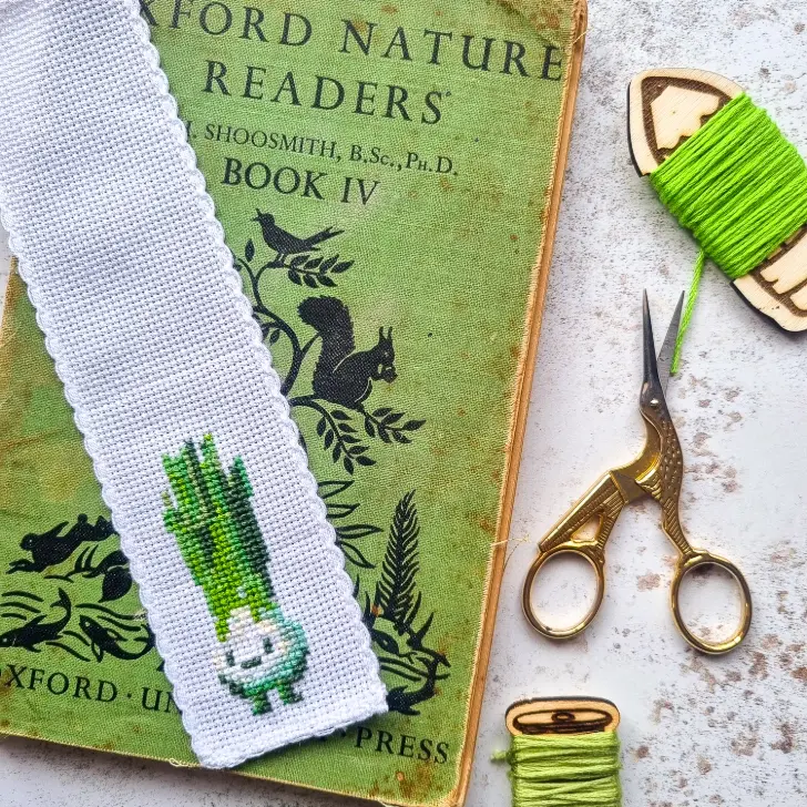 spring onion cross stitch bookmark on a green book with a pair of gold stork scissors and three lots of green embroidery thread
