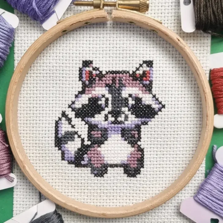 A wooden embroidery hoop holds a completed cross-stitch of a cute pixel-art-style baby raccoon on white Aida fabric. The raccoon is stitched in black, white, and shades of purple, with small red details in its ears. Surrounding the hoop are embroidery floss bobbins and skeins in coordinating colors, including purples, reds, and browns. The fabric is neatly stretched within the hoop, and the stitches are evenly placed, creating a cute handcrafted design.