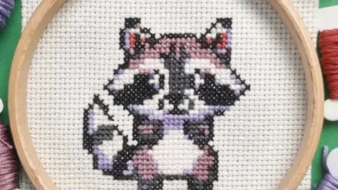 A wooden embroidery hoop holds a completed cross-stitch of a cute pixel-art-style baby raccoon on white Aida fabric. The raccoon is stitched in black, white, and shades of purple, with small red details in its ears. Surrounding the hoop are embroidery floss bobbins and skeins in coordinating colors, including purples, reds, and browns. The fabric is neatly stretched within the hoop, and the stitches are evenly placed, creating a cute handcrafted design.