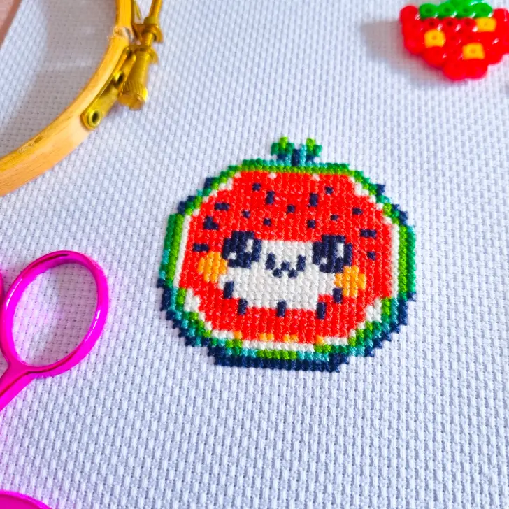 A cute watermelon slice cross stitch design on white Aida fabric, featuring a smiling face with rosy cheeks in a pixel art style. The stitched piece is surrounded by crafting tools, including pink embroidery scissors and a wooden embroidery hoop. A small strawberry-shaped beadwork decoration is visible in the background.