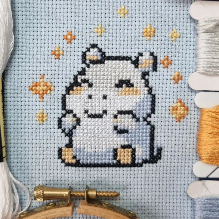 A cross-stitch pattern featuring a happy cartoon hippo in shades of grey and white, with orange and yellow stars scattered around on a light blue fabric background. The piece is displayed in a small embroidery hoop, with threads and bobbins in coordinating colors visible nearby.