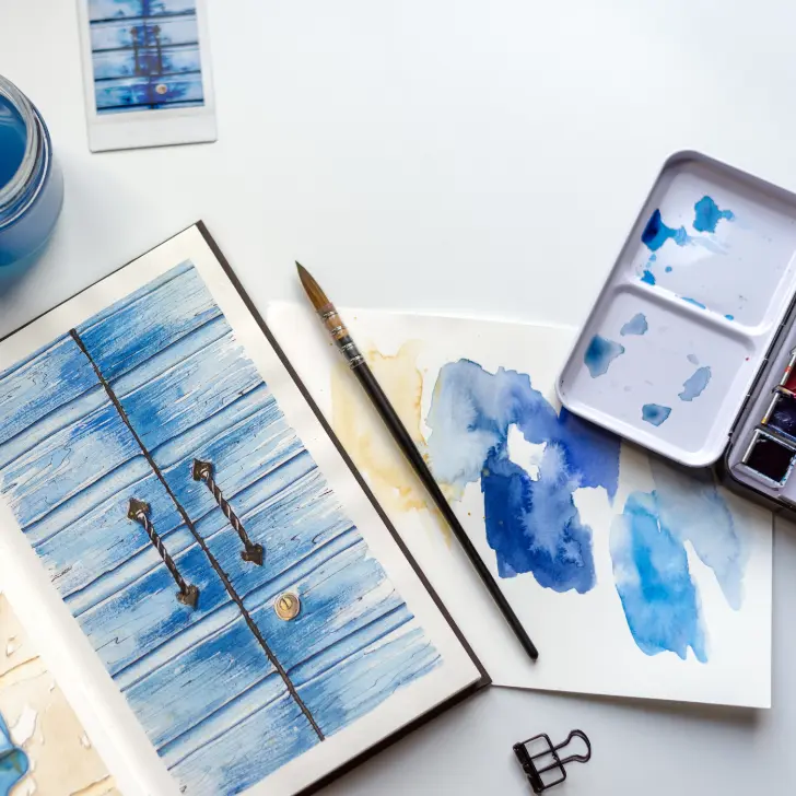 A flat lay of a watercolor painting setup. The centerpiece is an open sketchbook with a painting of a rustic blue wooden door, complete with detailed textures and handles. Surrounding the sketchbook are a paintbrush, a palette with shades of blue watercolor, a glass jar of water, and a sheet of paper with blue paint swatches. A small binder clip and a Polaroid photo of the door add to the artistic workspace aesthetic. The white background enhances the focus on the vibrant blue tones.