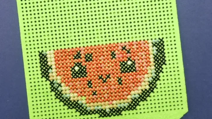 A bright green perforated felt banner with a cross-stitched design of a smiling watermelon slice. The watermelon has black seeds and a cheerful expression, surrounded by shades of red, pink, green, and white. The banner is threaded with a teal ribbon at the top for hanging, displayed on a dark blue background.