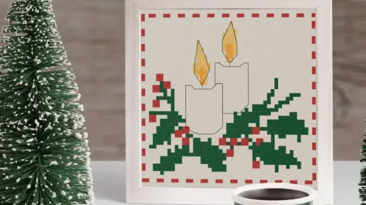 Image shows an embroidered chrsitsmas candle cross stitch framed in a white sqaure frame with two decorative small trees either side and a green mug in front.