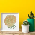 Images shows an embroidered yellow cross stitch rose, framed in a white square frame on a wooden table with two cacti in post next to it