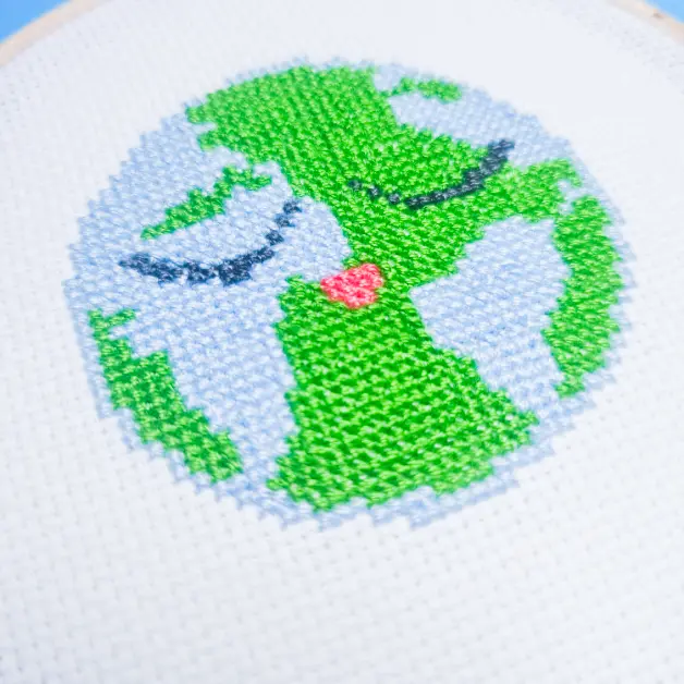 image shows embroidered planet earth cross stitch framed in a wooden hoop against blue and green paper background